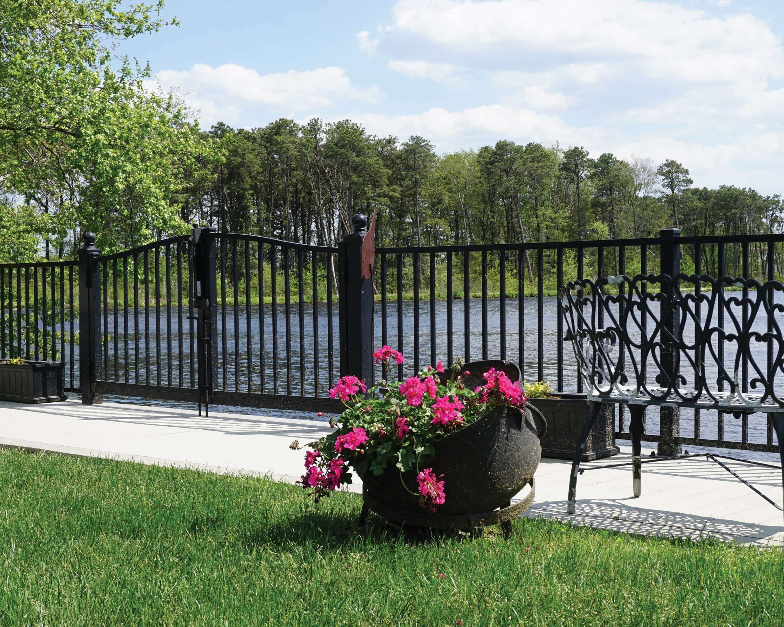 A large outdoor flower pot with striking pink flowers sits in front of an Active Yards Bedrock aluminum fence with Arched Gate between the viewer and the lake.